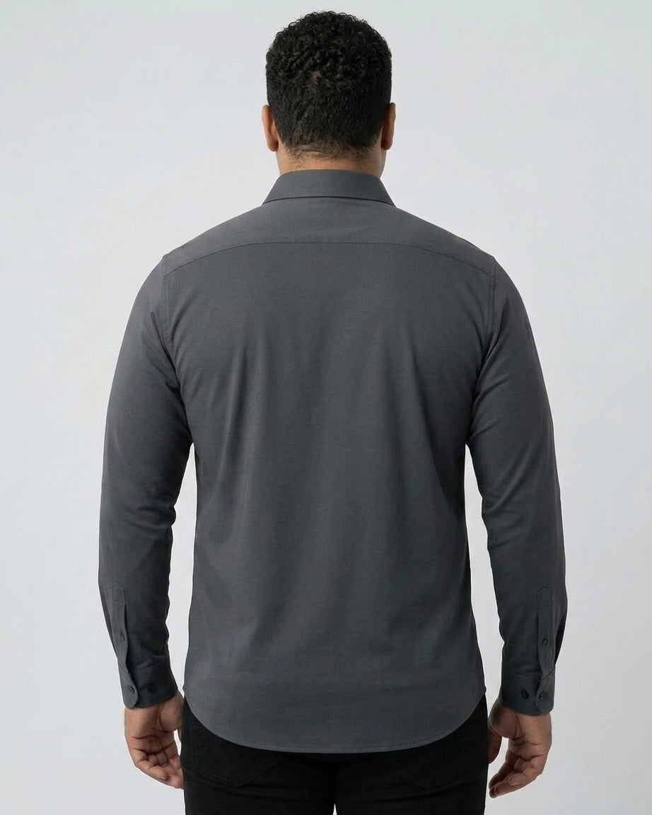 Homem de costas usando camisa social cinza-escuro de mangas longas. A imagem mostra o caimento da peça nas costas, a gola e a barra arredondada sobre uma calça preta.