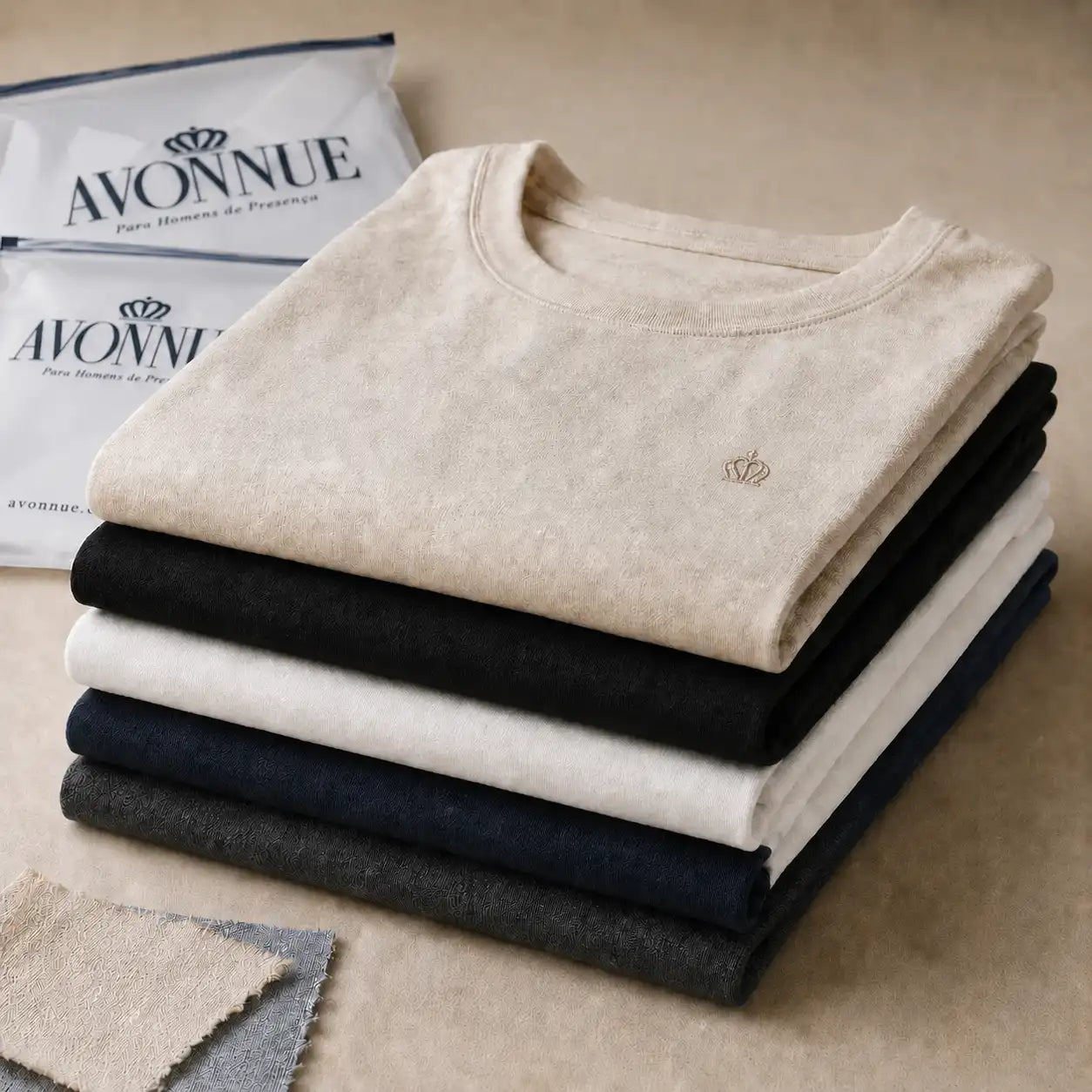 Imagem da secao Tecidos AVONNUE com camisetas masculinas dobradas em tons off white, preto, azul marinho e cinza sobre mesa.