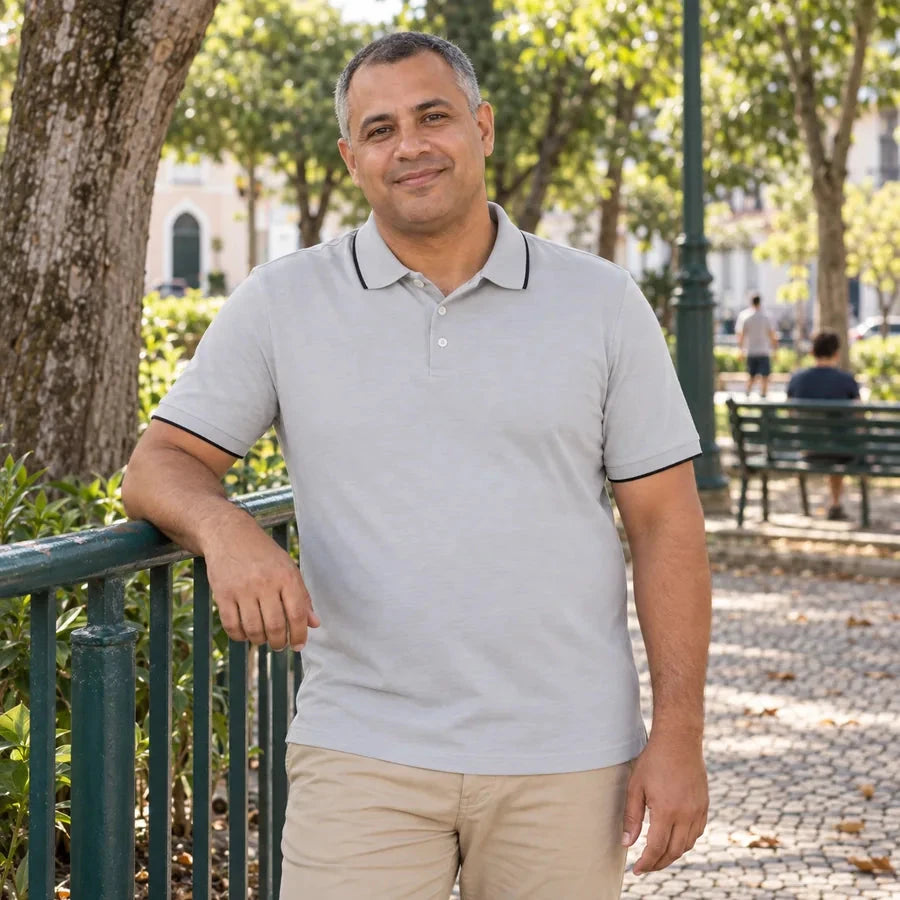 Homem usando polo cinza com detalhes escuros na gola e nas mangas, por fora da calça bege, apoiado em um guarda-corpo verde em uma praça arborizada.