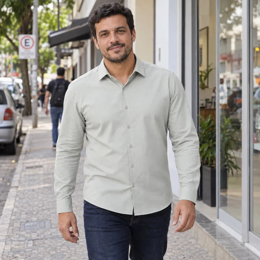 Homem caminhando em uma calçada comercial, usando camisa verde-clara de mangas longas por fora de um jeans azul-escuro.
