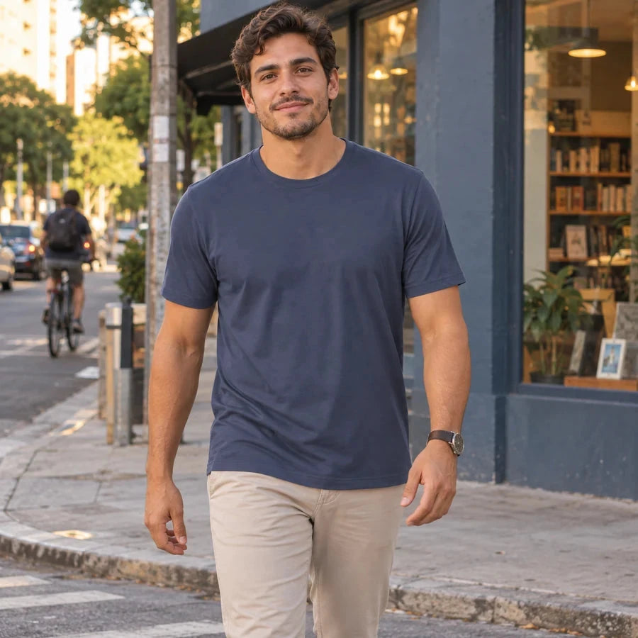 Homem jovem caminhando em uma rua ao fim da tarde, usando camiseta azul-marinho de mangas curtas por fora de uma calça bege-clara.