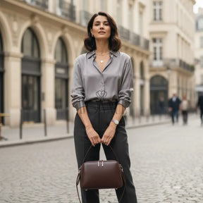 Modelo feminina AVONNUE em look cinza elegante, segurando bolsa marrom estruturada de mão em cenário urbano sofisticado.