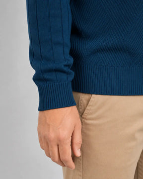Close da mão de um homem ao lado do corpo, mostrando a manga do suéter azul-marinho com linhas verticais, punho canelado ajustado ao pulso e barra canelada sobre uma calça bege.