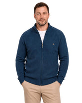 Homem de frente usando suéter azul-marinho com gola alta parcialmente aberta, zíper frontal inteiro e camiseta branca por baixo. A peça tem trama geométrica no peito, mangas com linhas verticais e barra canelada, combinada com calça bege.