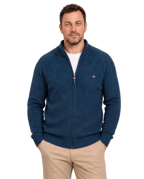 Homem de frente usando suéter azul-marinho com gola alta parcialmente aberta, zíper frontal inteiro e camiseta branca por baixo. A peça tem trama geométrica no peito, mangas com linhas verticais e barra canelada, combinada com calça bege.