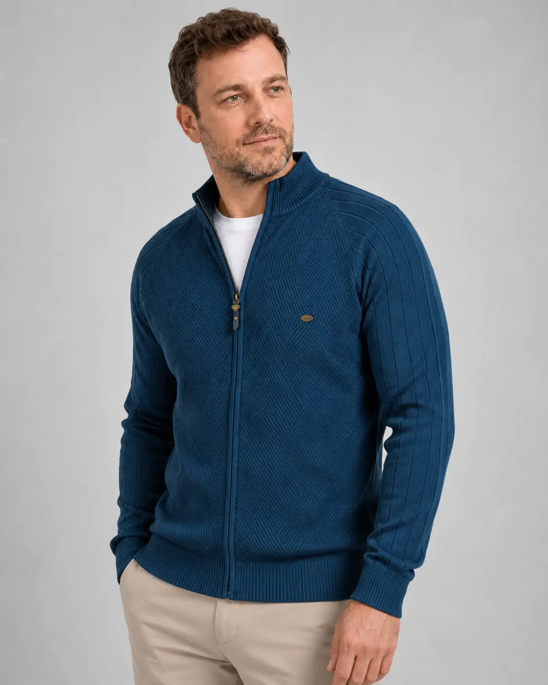 Homem em posição levemente lateral usando suéter azul-marinho com zíper frontal, gola alta parcialmente aberta e camiseta branca por baixo. A peça tem trama geométrica no tronco, mangas caneladas e barra ajustada, combinada com calça bege.