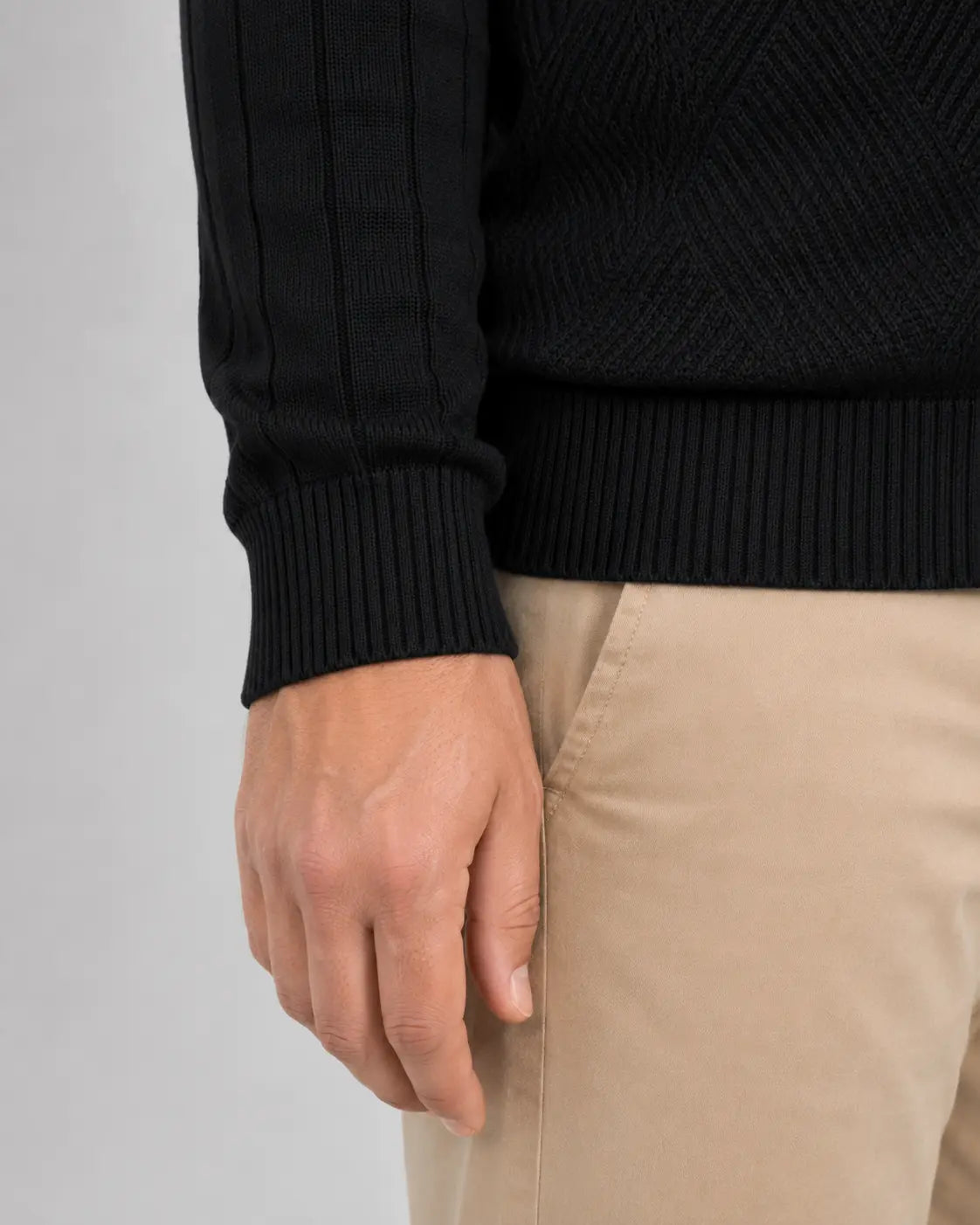 Close da manga do suéter preto, mostrando o punho canelado ajustado ao pulso, a barra canelada e parte da trama geométrica frontal. O homem usa calça bege.
