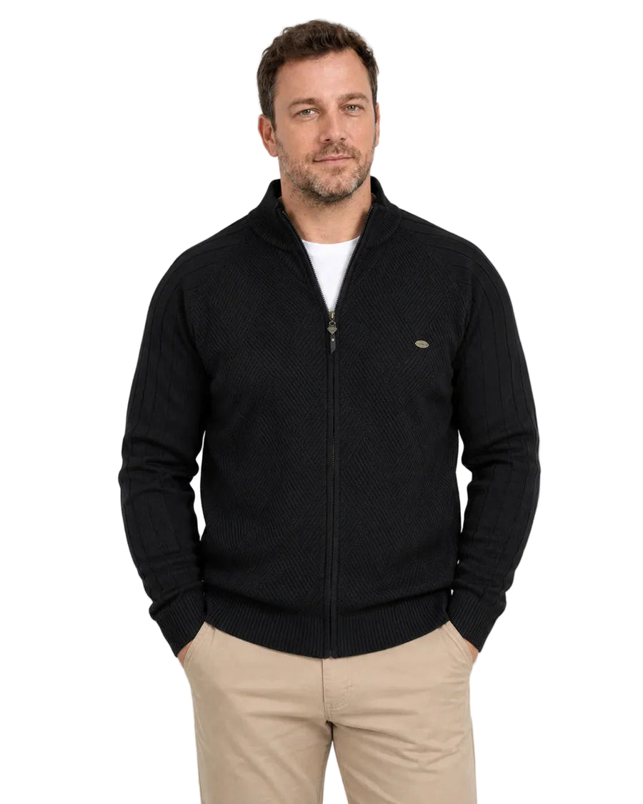 Homem de frente usando suéter preto com zíper frontal inteiro, gola alta e camiseta branca aparecendo no decote. A peça tem trama geométrica no peito, mangas caneladas e barra ajustada, combinada com calça bege.