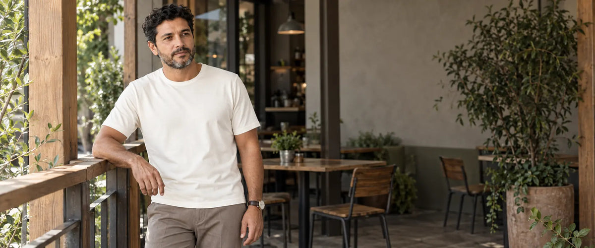 Homem usando camiseta off-white de mangas curtas e calça bege acinzentada, apoiado em um guarda-corpo de madeira na varanda de um café.
