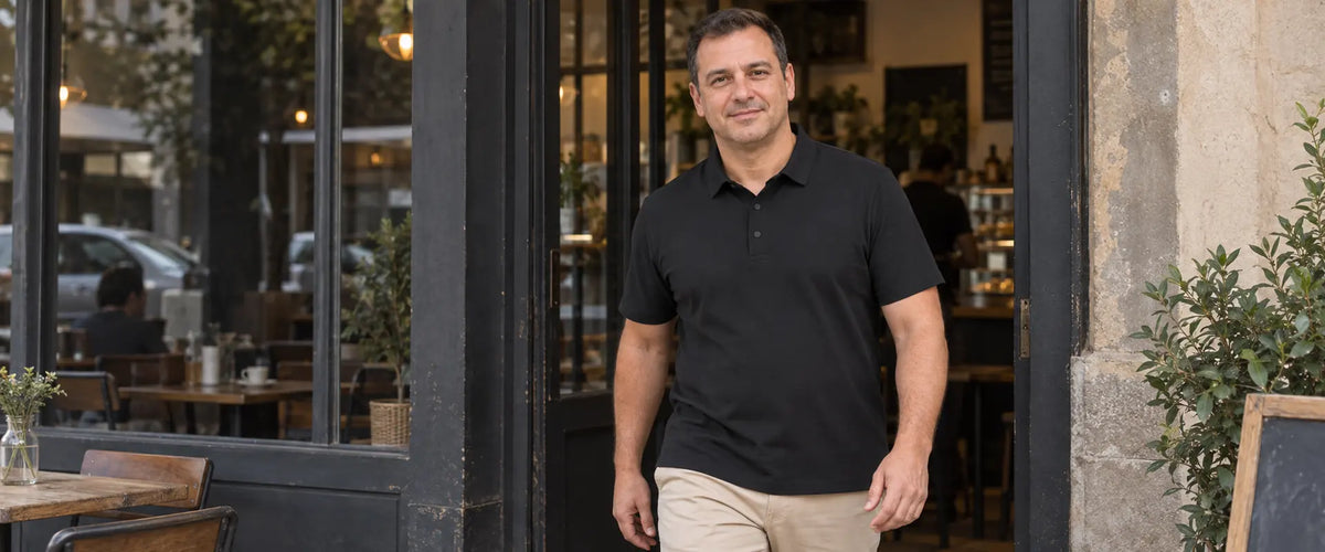 Homem na entrada de um café, usando polo preta de mangas curtas por fora da calça bege, com os braços relaxados e as mãos visíveis.